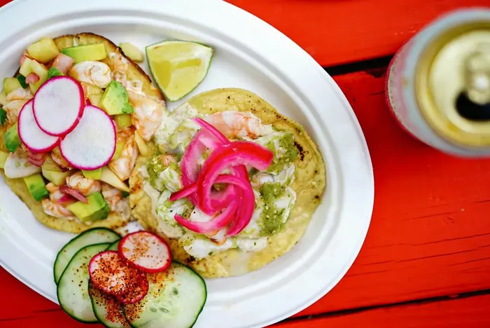 Two shrimp tostadas with avocado, sliced radish and pink pickled onion on a white plate, on a red table