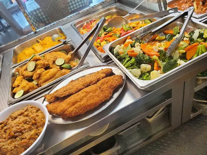 A steam table of fried fish fillets, stewed beans, broccoli and carrots, with serving tongs